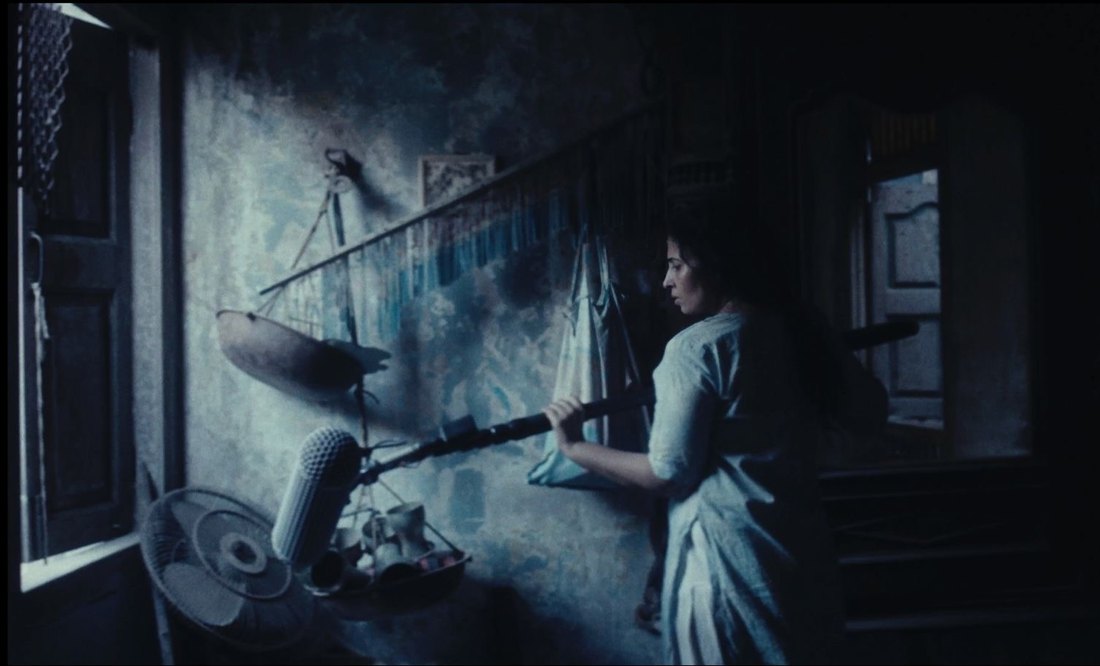 Film still: a woman holds a boom microphone against a bare plaster wall in a dim interior.