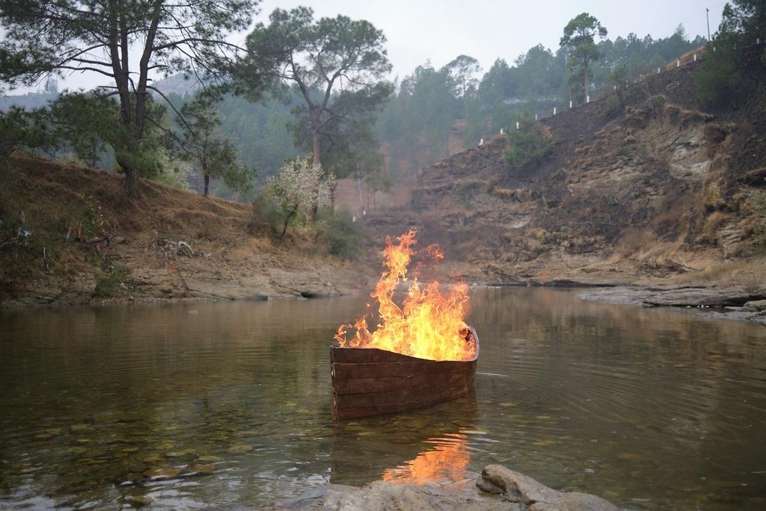 A burning boat on the Kumaon river below a bare hillside.